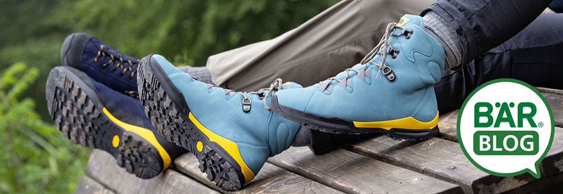 Close-up of two pairs of hiking boots, one blue and one dark, resting on a wooden bench with a green outdoor background.