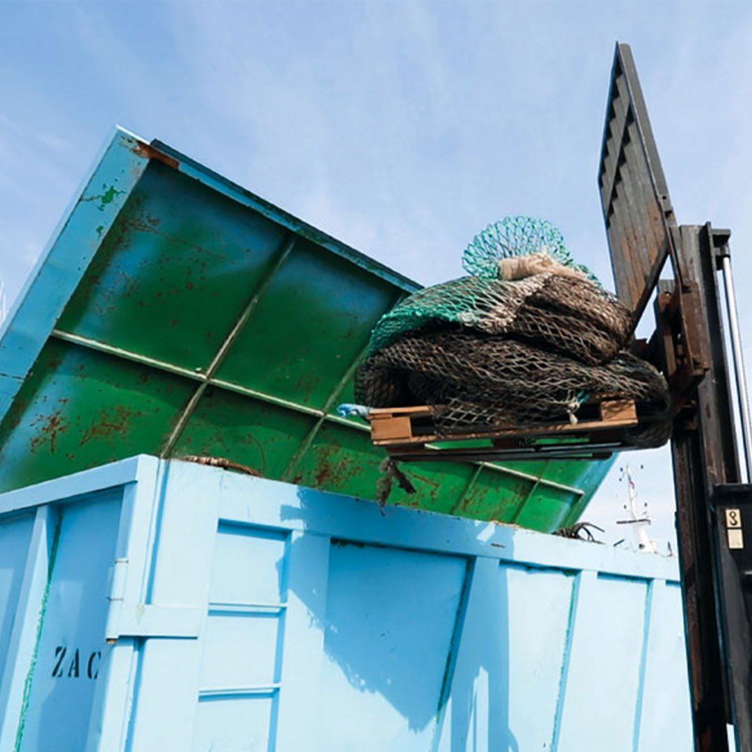 Gabelstapler entlädt große Netze in blauen Container Ein Gabelstapler hebt ein Bündel alter Fischernetze in einen großen, offenen blauen Container, der im Freien steht.