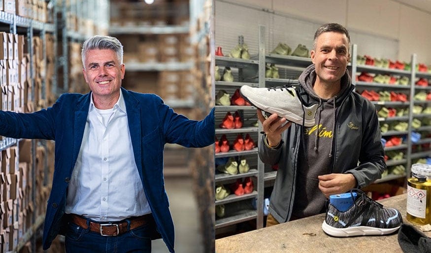 Zwei Männer in einem Lager mit Schuhen: links ein Mann in einem blauen Anzug vor Regalen mit Kartons, rechts ein Mann in dunkler Kleidung hält einen Schuh. Links ein Mann in einem Anzug in einem Lager mit Kartons, rechts ein Mann in dunkler Kleidung mit einem Schuh in der Hand, im Hintergrund Regale mit Schuhen.