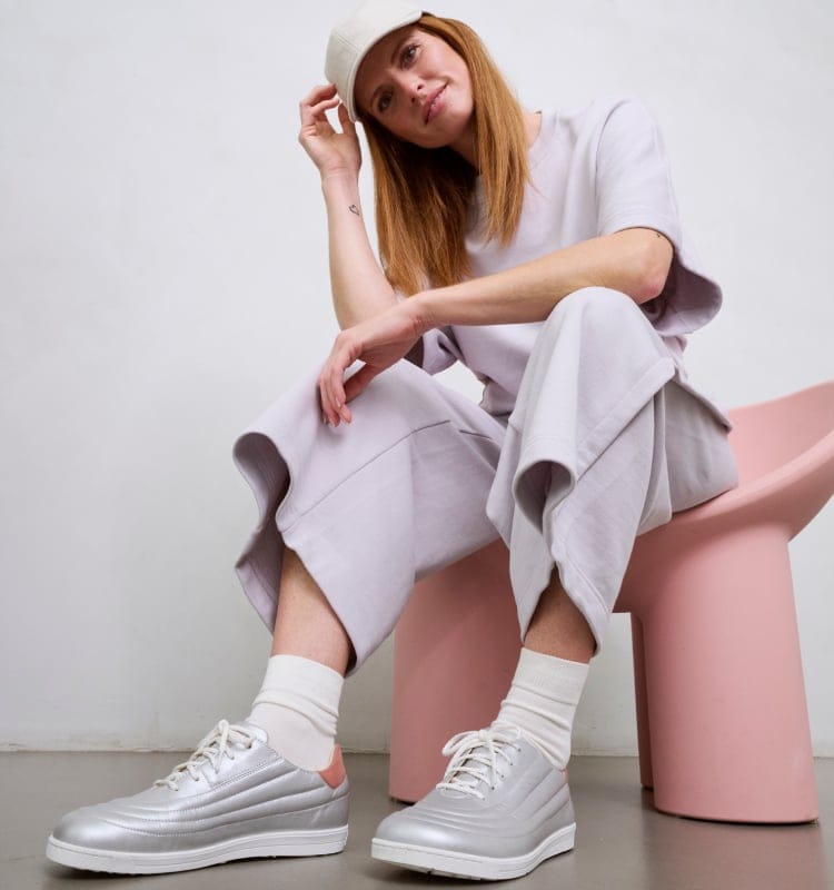 Frau in hellem Outfit und silbernen Sneakers auf rosa Stuhl sitzend