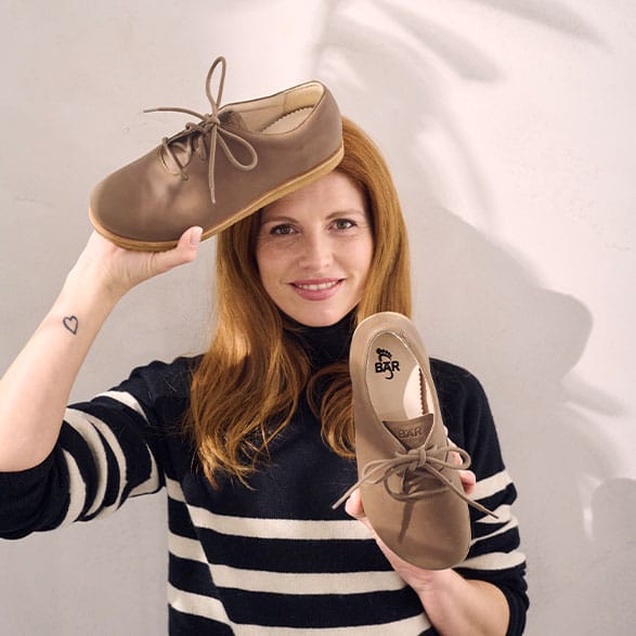 A smiling woman with red hair holding up a pair of beige lace-up shoes, showcasing their design and style