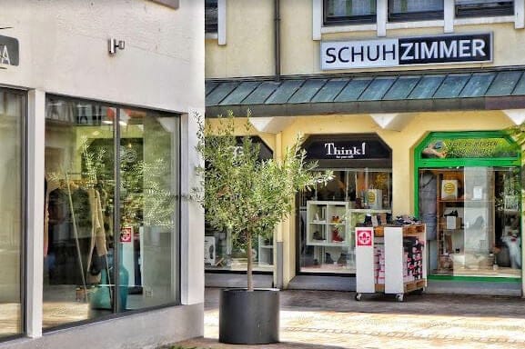 Eingangsbereich eines Schuhgeschäfts im Freien Foto eines kleinen Einkaufsbereichs mit Schaufenstern und dem Eingang eines Schuhgeschäfts namens Schuhzimmer. Es gibt einen Olivenbaum im Topf vor dem Geschäft.
