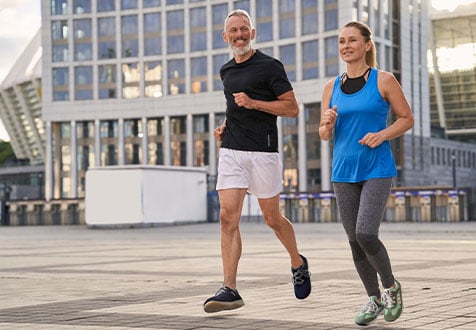 Zwei Personen joggen in sportlicher Kleidung durch eine urbane Umgebung mit modernen Gebäuden im Hintergrund.