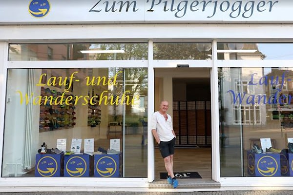 Schaufenster des 'Zum Pilgerjogger' mit Lauf- und Wanderschuhen, Mann steht in der Tür