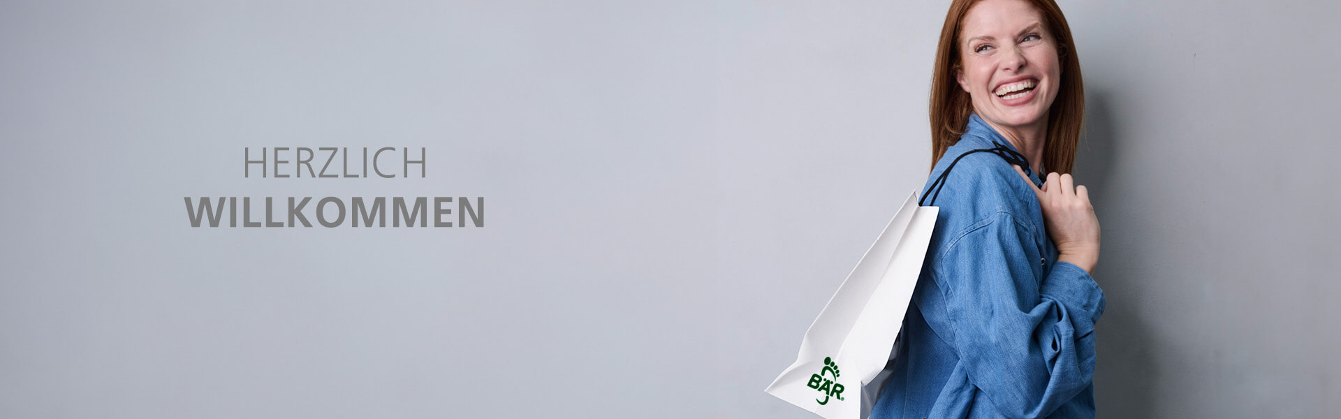 Fröhliche Frau mit Einkaufstüte willkommen im Shop Lächelnde Frau in Jeanshemd trägt eine Einkaufstasche. Text: Herzlich Willkommen.