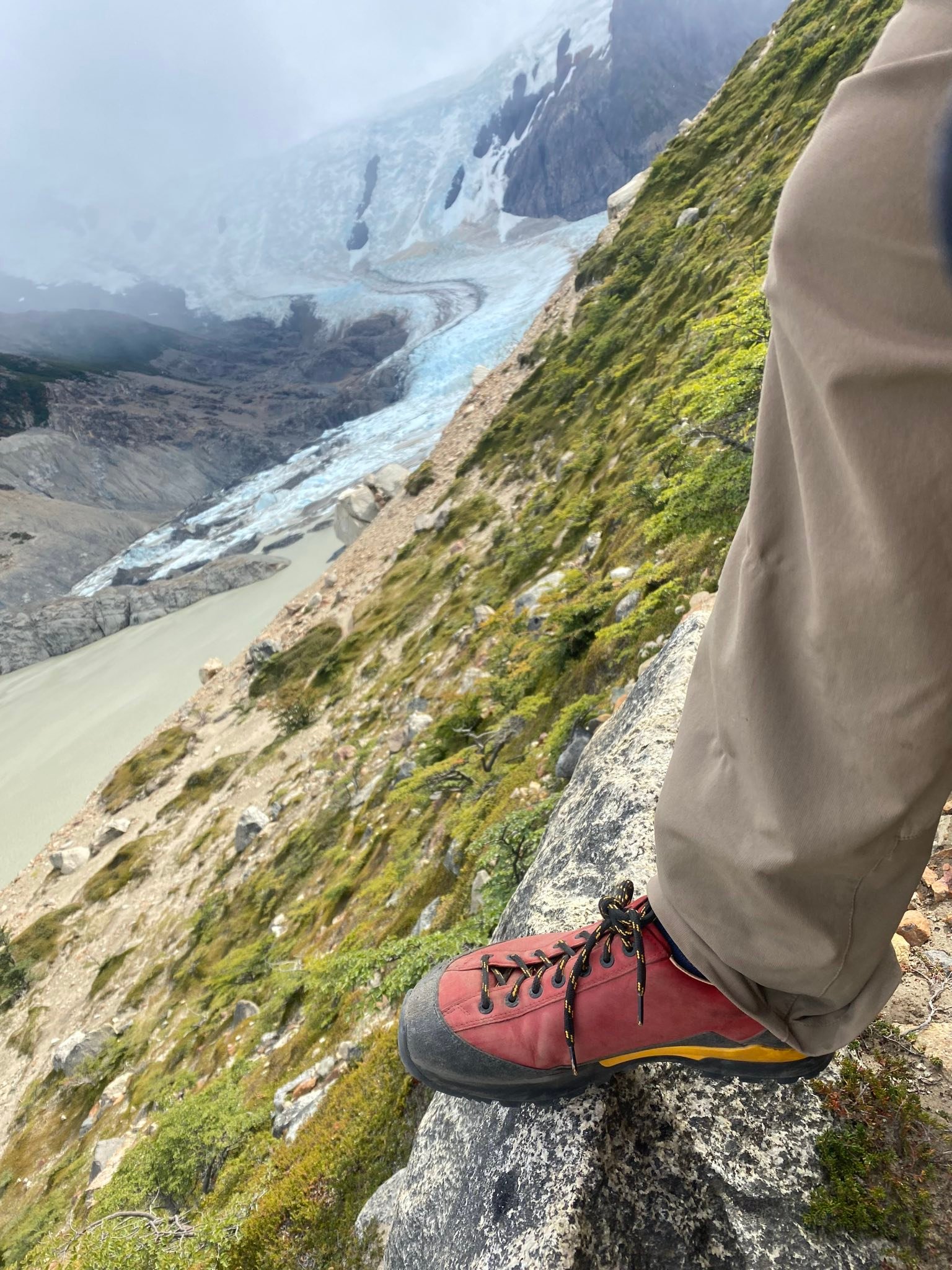 Ein roter Wanderschuh steht auf einem Felsen mit Blick auf einen Gletscher und eine bergige Landschaft im Hintergrund.