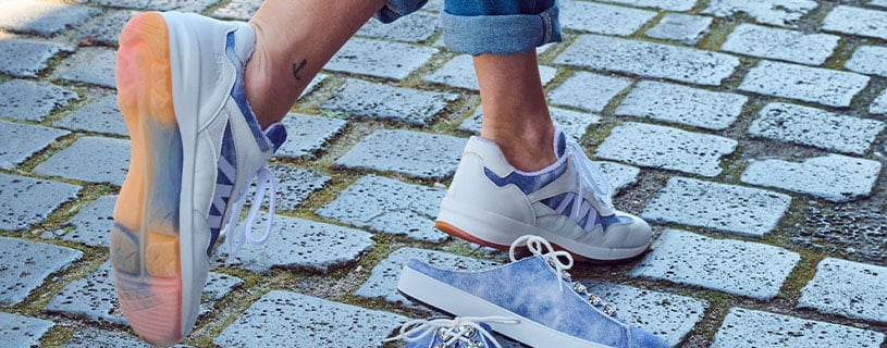 Person mit modischen Sneakern geht auf Kopfsteinpflaster, ein weiteres Paar Sneaker liegt daneben