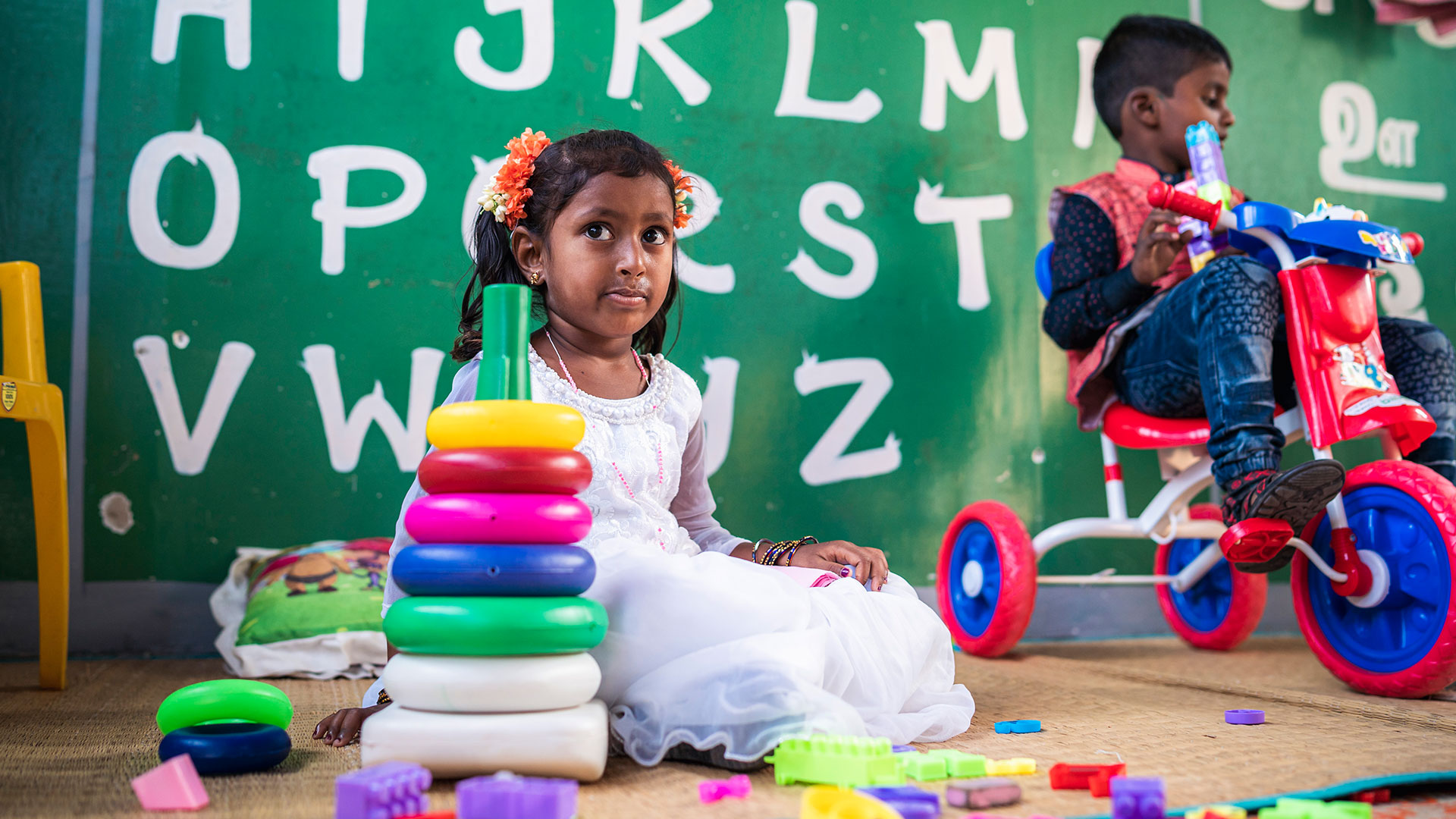 Kind beim Spielen im Kindergarten mit buntem Spielzeug Ein Kind sitzt im Kindergarten auf dem Boden und spielt mit buntem Spielzeug. Im Hintergrund ist eine grüne Wand mit aufgemalten Buchstaben.