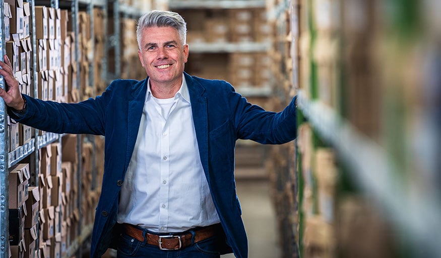 Confident businessman standing in a warehouse aisle Smiling man in a blue jacket and white shirt in a warehouse setting, surrounded by shelves and boxes