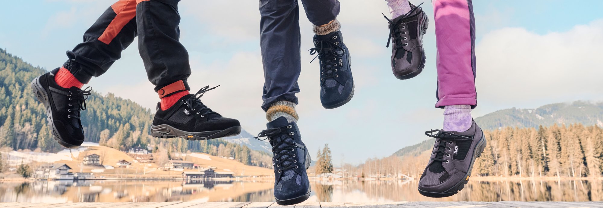 Sprung im Freien mit Outdoor-Schuhen vor malerischer Landschaft Füße in Outdoor-Schuhen springen in die Luft, vor einer malerischen Landschaft mit Bergen und einem See im Hintergrund.