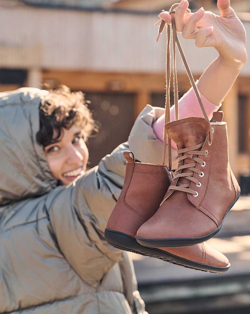 Lachende Frau in Winterjacke hält braune Schnürstiefel hoch