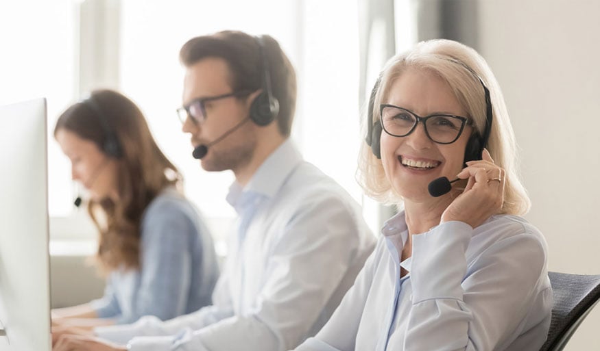 Freundliches Kundensupport-Team mit Headsets bei der Arbeit Drei Personen im Kundensupport mit Headsets, die an Computern arbeiten