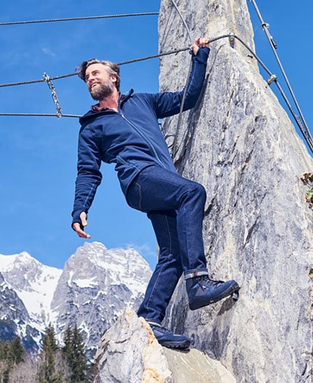 Mann in Outdoor-Bekleidung beim Klettern in den Bergen Mann mit blauer Jacke und Jeans klettert auf einem Felsvorsprung in einer Berglandschaft.