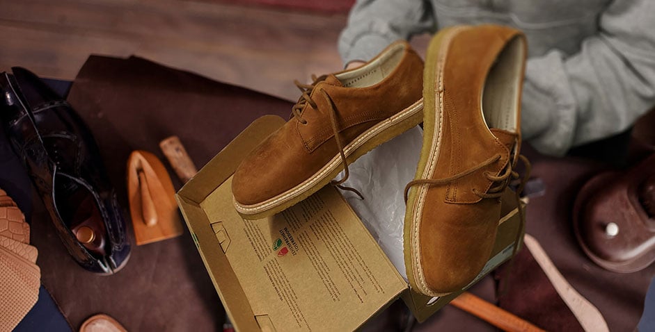 A pair of brown suede shoes being unboxed, surrounded by various footwear and accessories on a wooden table.