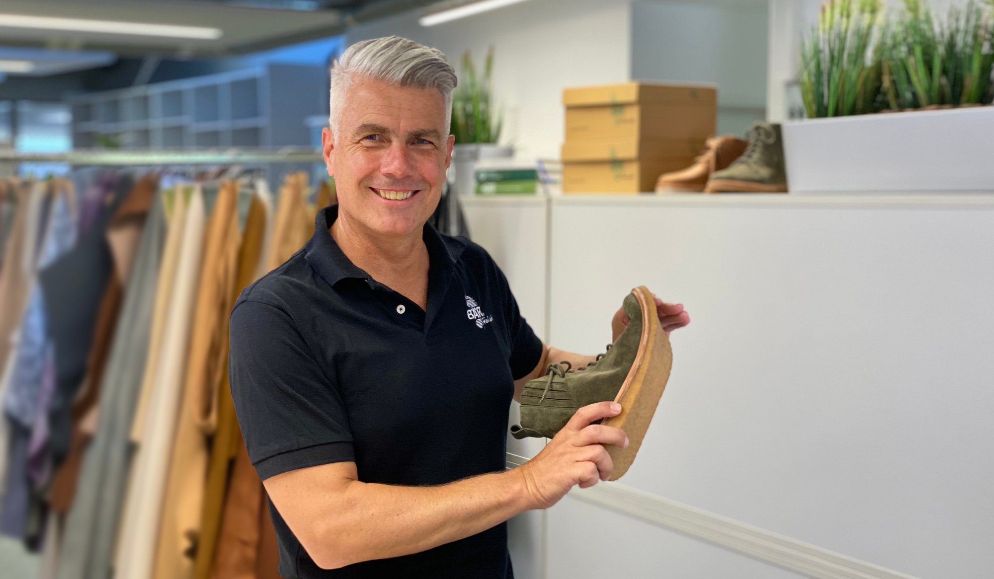 Mann in schwarzem Polo zeigt grünen Schuh in Büro Mann mit grauen Haaren in schwarzem Polohemd hält grünen Schuh im Büro mit Kleiderständern im Hintergrund