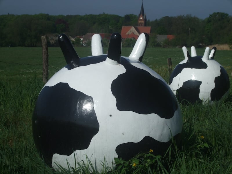Zwei große, kugelförmige Skulpturen in schwarz-weißer Kuhoptik auf einer grünen Wiese vor einem Dorf mit Kirchturm.