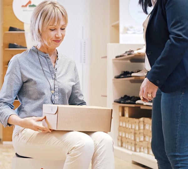 A woman sitting down holding a shoe box, assisted by another person in a shoe store.