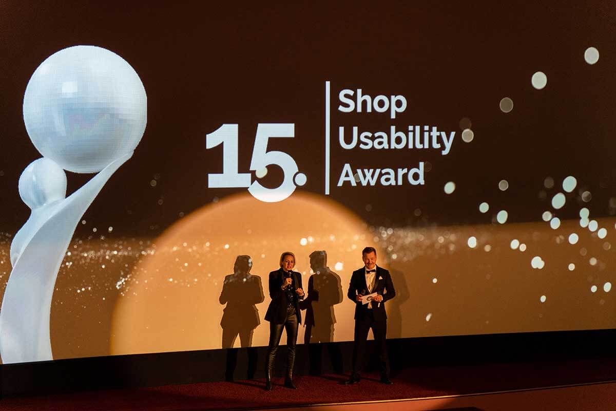 15. Shop Usability Award Verleihung mit Moderatoren auf der Bühne Moderatoren stehen auf der Bühne bei der 15. Verleihung des Shop Usability Awards, mit Award-Logo im Hintergrund.