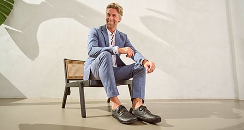 Stylish man in blue suit sitting on a chair in a bright room Man in blue suit and leather shoes sitting on a chair, casting shadow on white wall