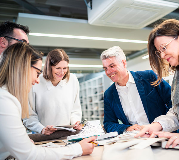 Teammeeting im Büro mit Fokus auf Design und Zusammenarbeit Eine Gruppe von Mitarbeitenden in einem Büro, die über einem Tisch voller Designunterlagen diskutieren und gemeinsam an einem Projekt arbeiten.