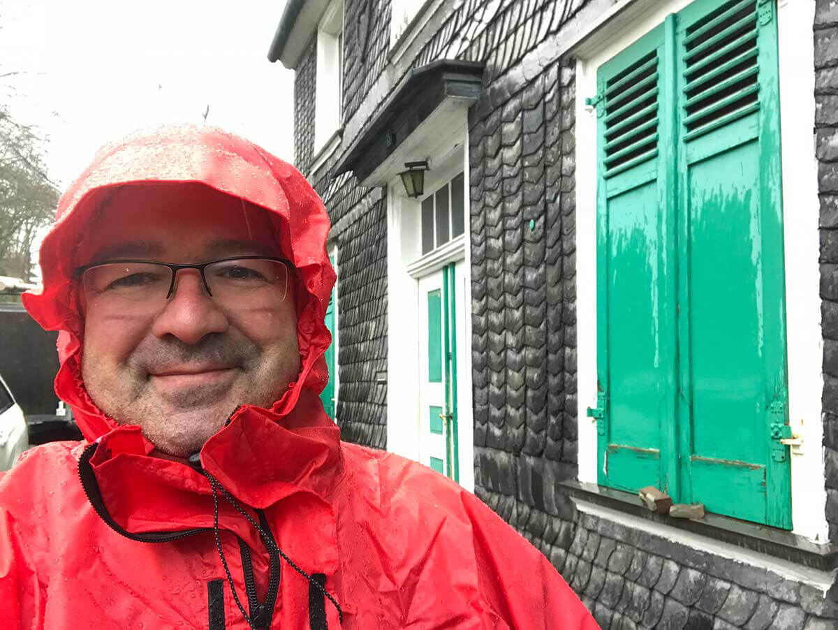 Man wearing glasses and a red raincoat smiling in front of a grey building with green shutters on a rainy day