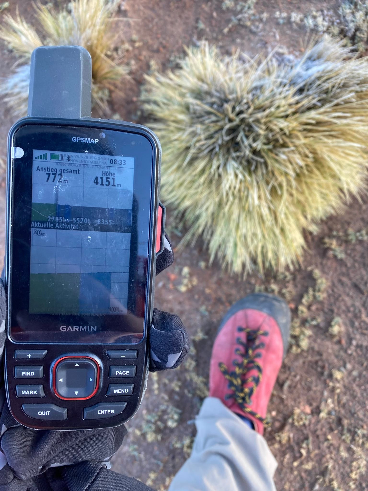 Hand hält GPS-Gerät, das eine Höhe von 4151 Metern anzeigt; im Hintergrund Bergsteigerschuh auf gebirgigem Gelände