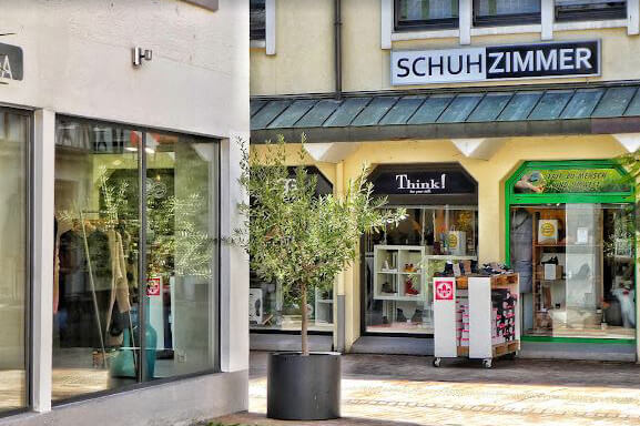 Eingangsbereich eines Schuhgeschäfts im Freien Foto eines kleinen Einkaufsbereichs mit Schaufenstern und dem Eingang eines Schuhgeschäfts namens Schuhzimmer. Es gibt einen Olivenbaum im Topf vor dem Geschäft.