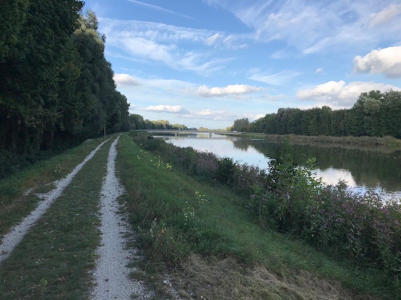 Gravelweg neben einem ruhigen Fluss, umgeben von Bäumen und blühenden Pflanzen unter einem blauen Himmel mit weißen Wolken