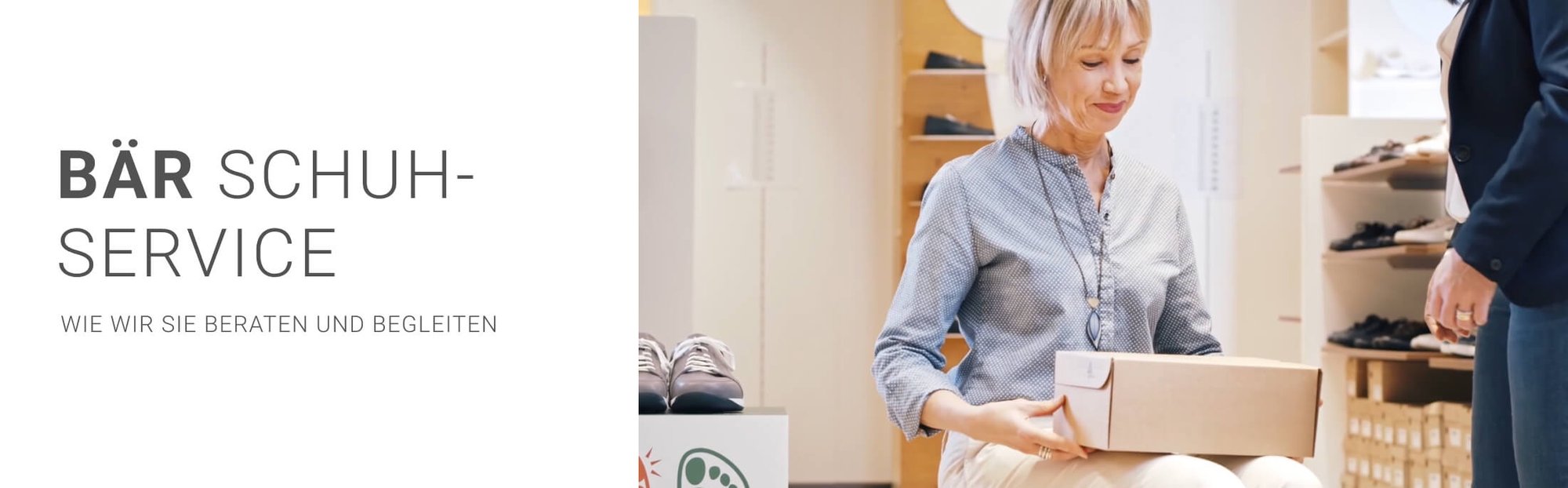 Frau sitzt in einem Schuhgeschäft und hält eine Schuhschachtel, während eine andere Person ihr hilft