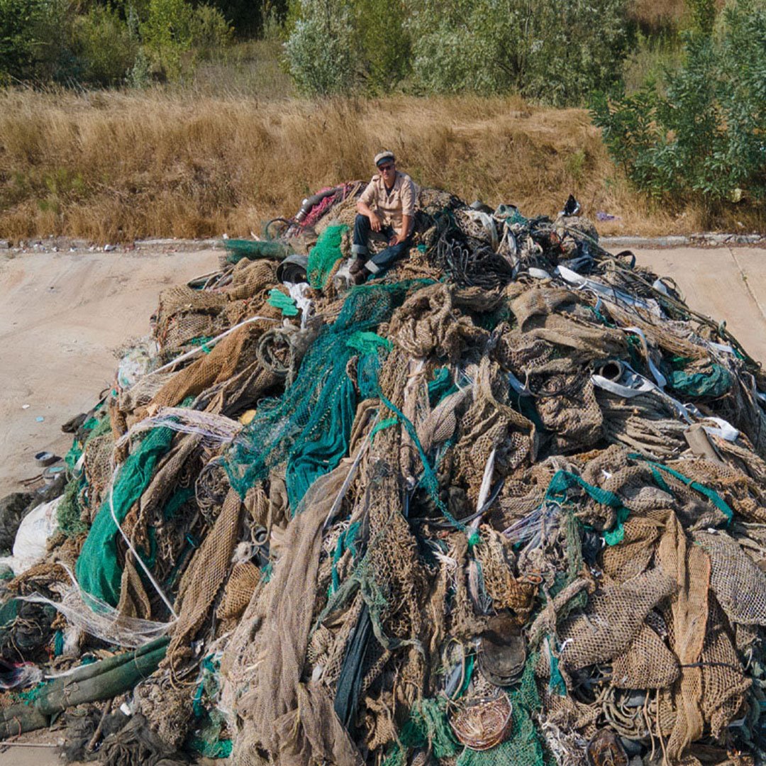 Person sitting on a large pile of discarded fishing nets outdoors A person is sitting on top of a huge pile of old, tangled fishing nets in an open area with grassy surroundings.