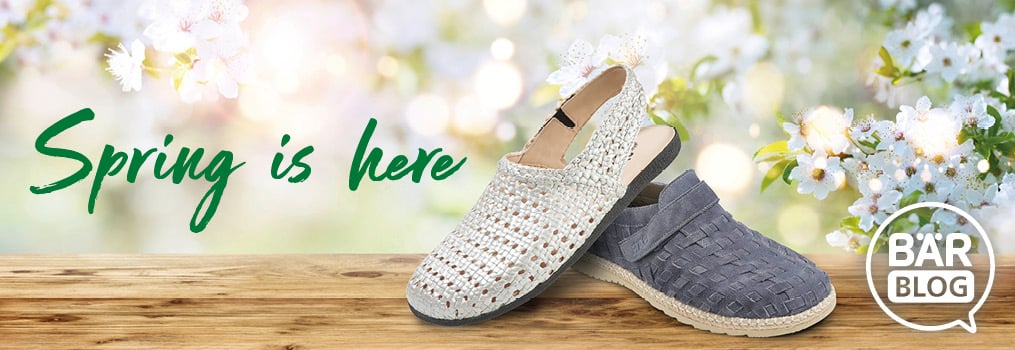 Comfortable Spring Slip-On Shoes for Women on a Blossoming Background Two pairs of woven slip-on shoes on a wooden surface with blossom flowers and 'Spring is here' text in the background