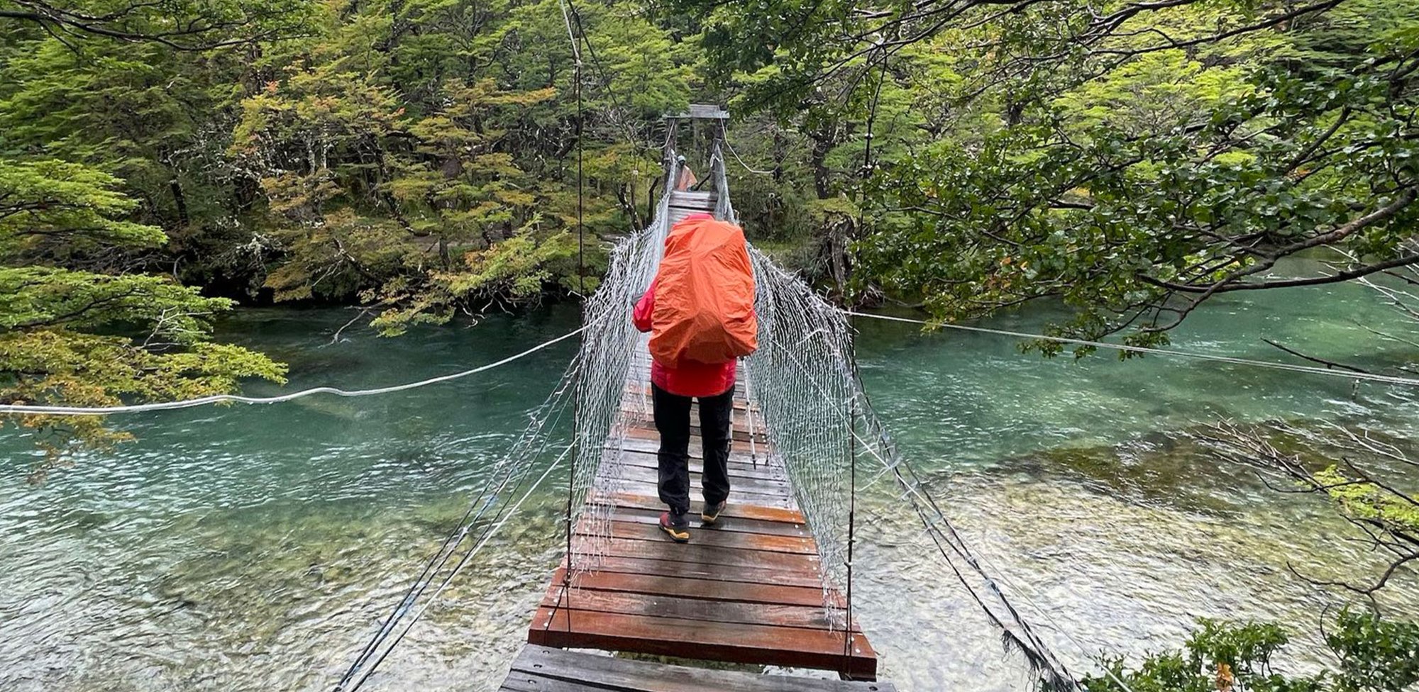 Wanderer mit orangem Rucksack auf Hängebrücke im Wald Person mit orangem Rucksack überquert eine Hängebrücke über einen klaren Fluss, umgeben von grünem Wald.