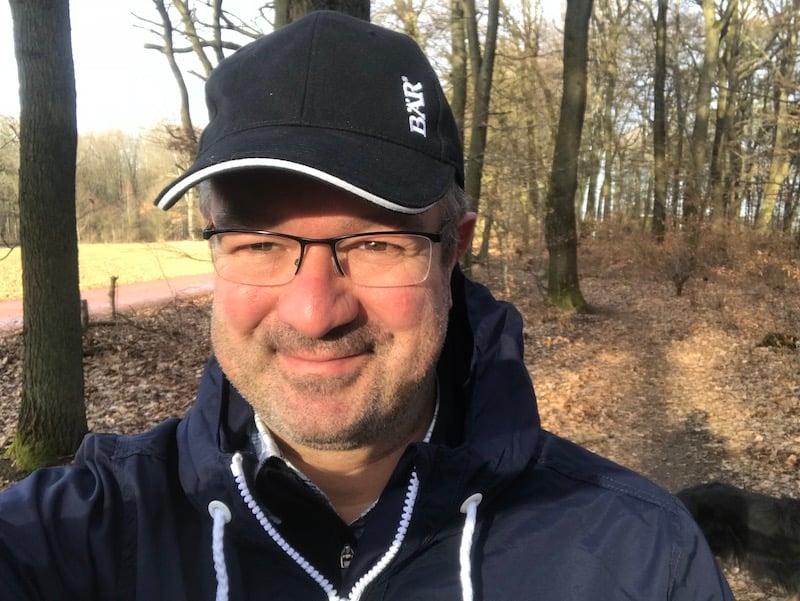 Ein Mann mit Brille und schwarzer Kappe steht lächelnd in einem sonnigen Wald.