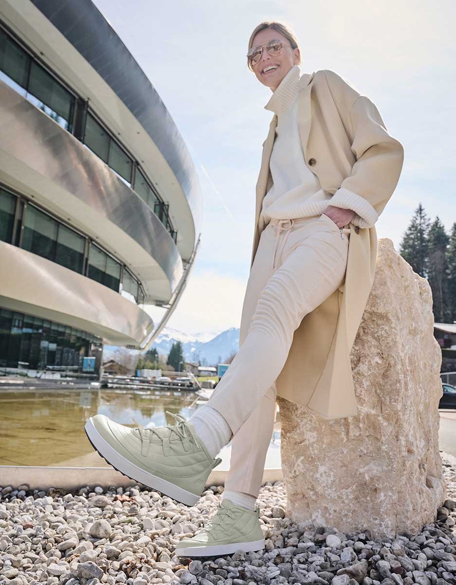 Person in beiger Kleidung und Sneakers lehnt l&auml;ssig an einem Stein, im Hintergrund modernes Geb&auml;ude und Berglandschaft