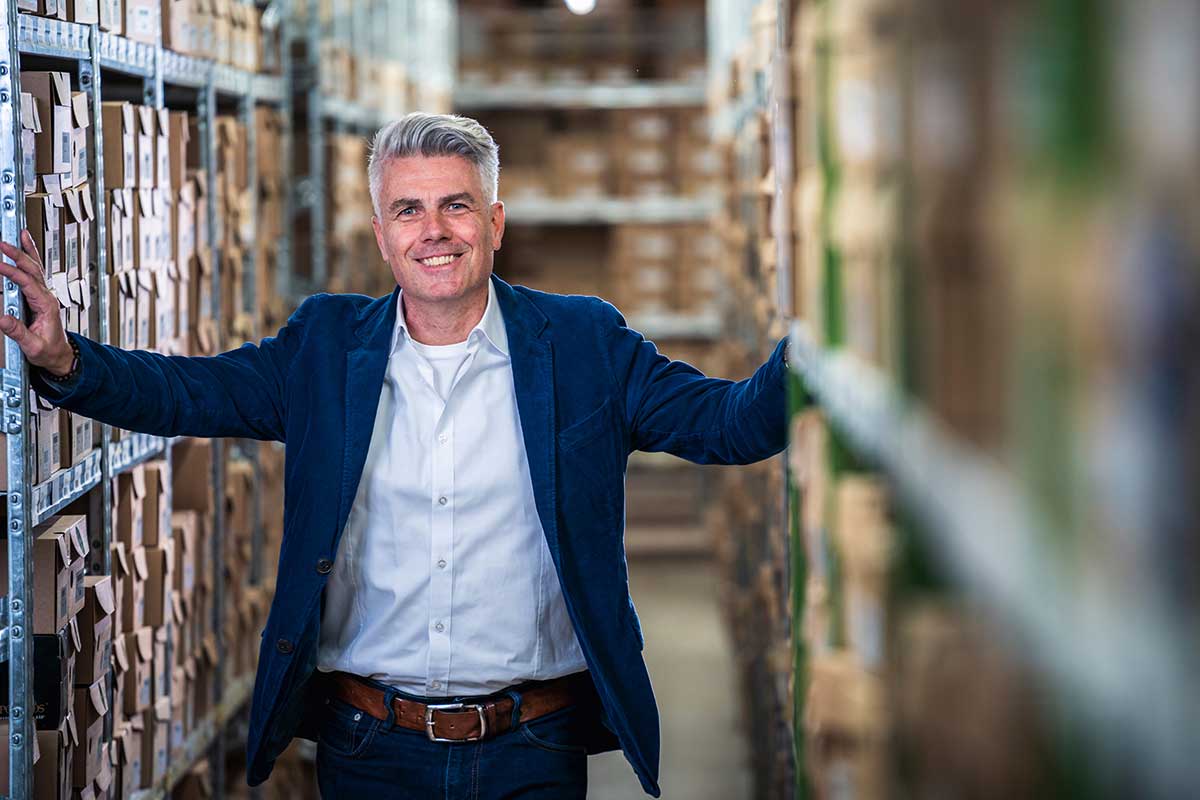 Businessman smiling in warehouse aisle with boxes on shelves Confident businessman in a blue blazer and white shirt standing in a warehouse aisle between shelves filled with boxes, smiling at the camera