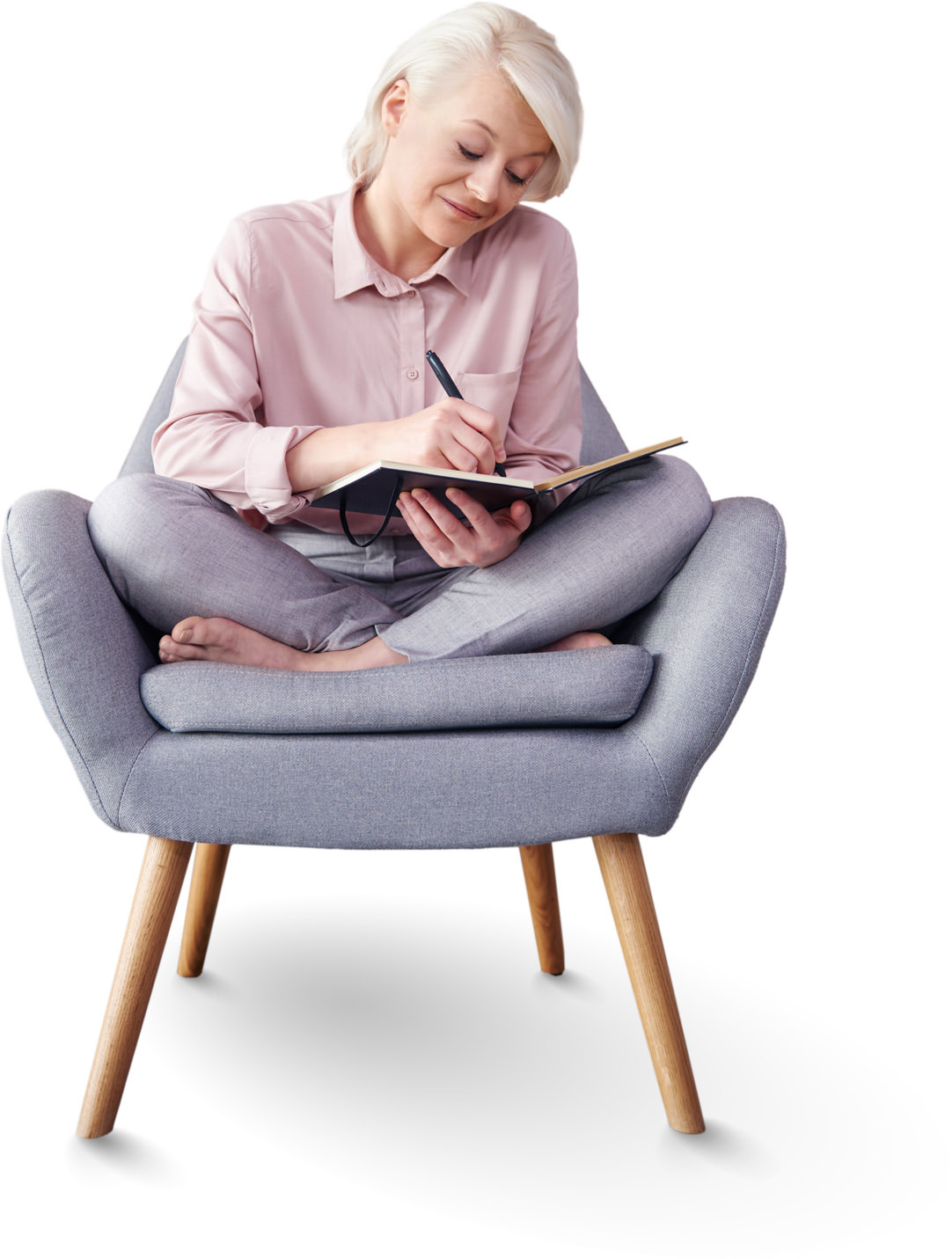 Woman in pink shirt writing in notebook while sitting on gray armchair Woman writing in a notebook on a gray cushioned armchair