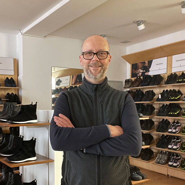 Freundlicher Verkäufer in einem Schuhgeschäft Ein lächelnder Mann steht mit verschränkten Armen in einem Schuhgeschäft. Im Hintergrund sind Regale mit verschiedenen Sportschuhen zu sehen.
