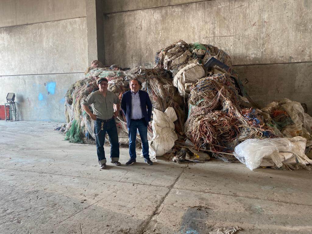 Two men standing beside a large pile of recyclable materials in an industrial warehouse setting.
