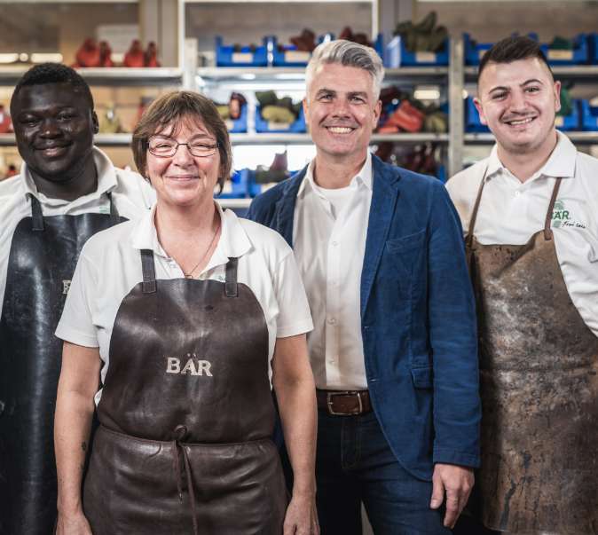 Team of Skilled Shoemakers in Workshop A group of four shoemakers, including three men and one woman, posed in a workshop setting, wearing aprons and smiling.