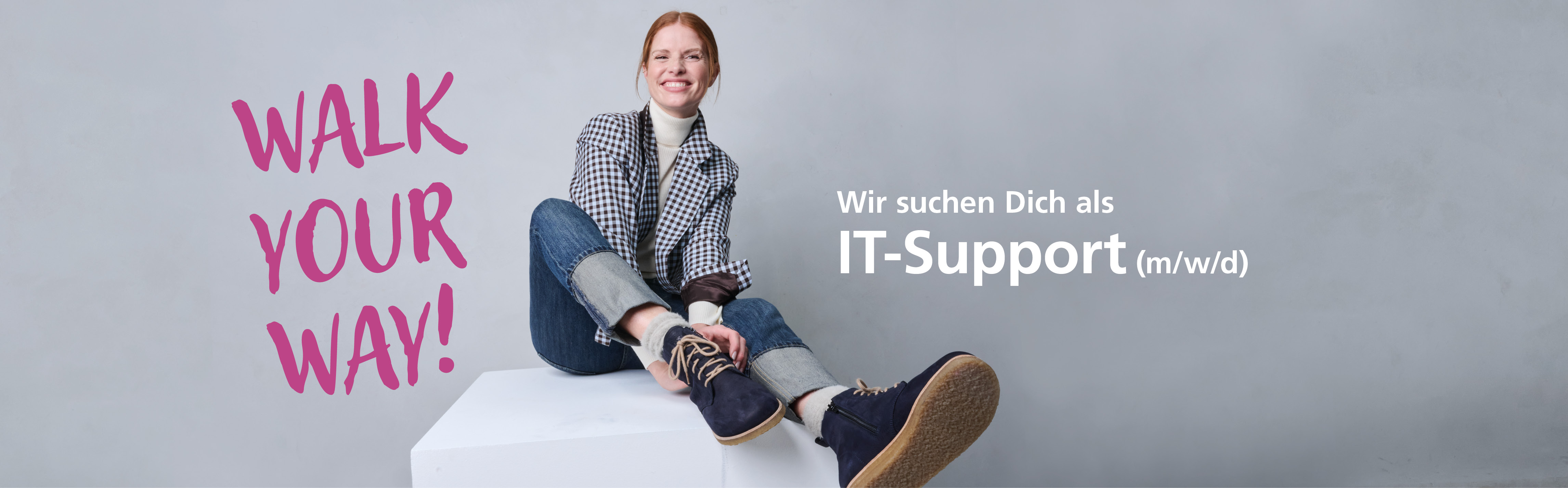 Person sitzt lächelnd auf einem Podest mit Text 'Walk your way! Wir suchen Dich als IT-Support (m/w/d)' auf grauem Hintergrund.