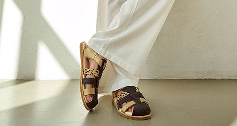 Modische Sandalen aus Leder für Damen - Sommerkollektion Damenfüße in stilvollen, braun-beigen Ledersandalen, weiße Hose, modernes Design