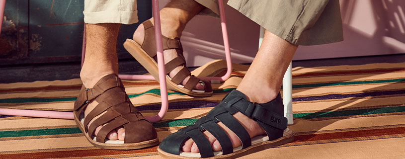 Zwei Personen tragen braune und schwarze Herrensandalen auf einem gestreiften Teppich