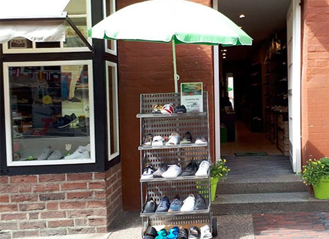 Schuhständer vor einem Laden mit Sonnenschirm Ein Schuhständer vor einem Geschäftseingang, geschützt von einem grünen Sonnenschirm, mit verschiedenen Sportschuhen ausgestellt.