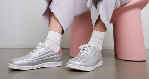 Stylish Silver Sneakers for Casual Wear Person wearing silver sneakers with white socks and light gray pants seated on a pink stool
