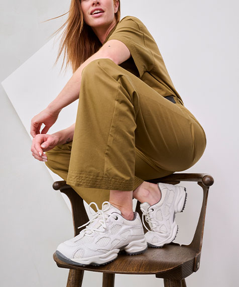 Stylish woman in khaki outfit with white sneakers seated on a wooden chair, showcasing a modern casual look