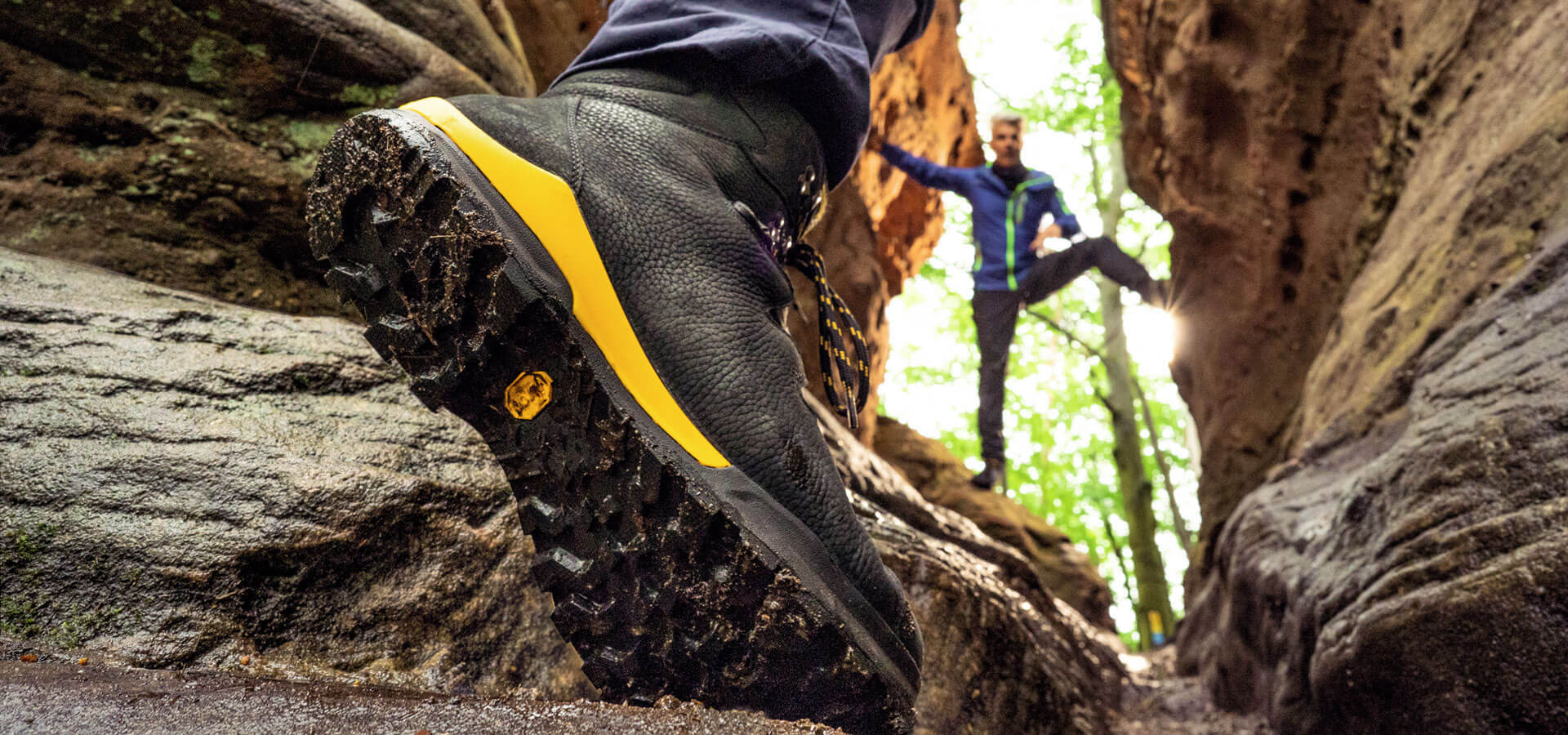 Robuste Wanderschuhe für anspruchsvolle Gelände Nahaufnahme eines robusten Wanderschuhs auf felsigem Untergrund mit Person im Hintergrund, die durch eine felsige Landschaft wandert