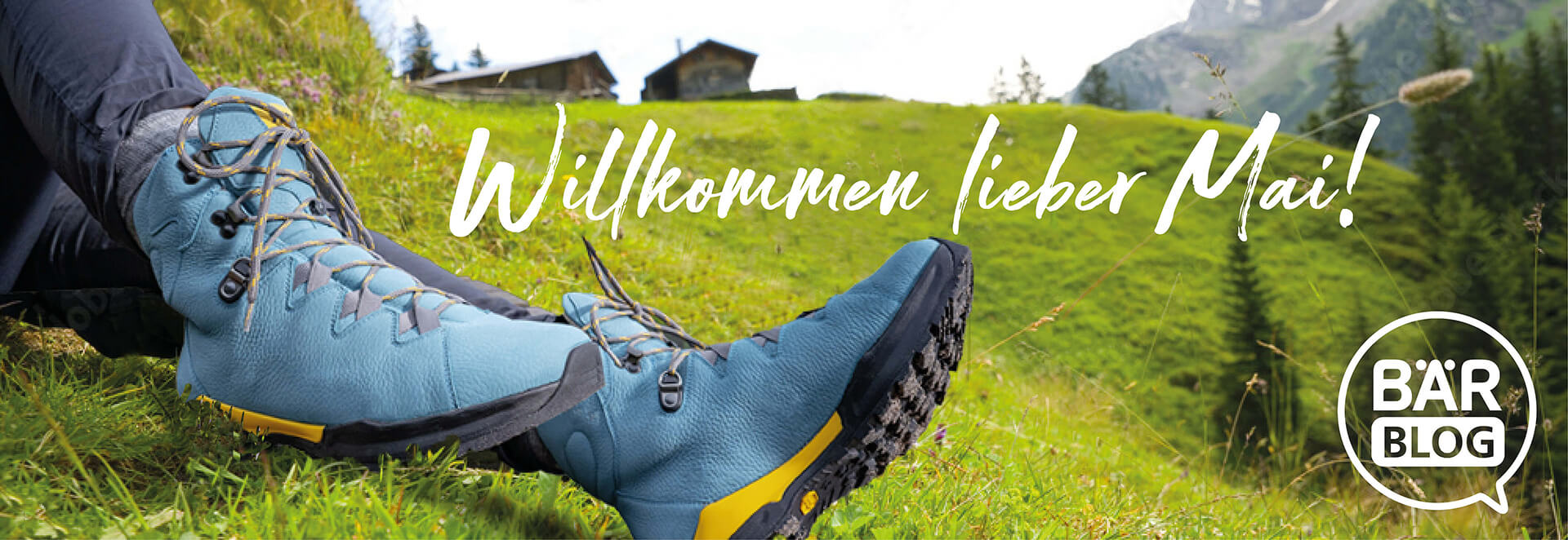 Hiking boots on a grassy hill welcoming May Close-up of blue hiking boots on a grass field with mountains in the background, text says 'Welcome dear May'