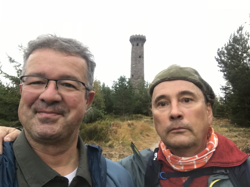 Two men outdoors, with one smiling and wearing glasses, standing near a stone tower surrounded by trees.