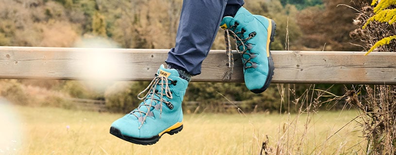 Outdoor hiking boots on a fence in scenic nature Person wearing blue hiking boots while sitting on a wooden fence in a grassy outdoor setting