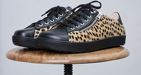 Trendige Sneaker mit Leopardenmuster und schwarzen Details Leopardenmuster Sneaker mit schwarzen Zehenkappen und Schnürsenkeln auf einem runden Holzhocker